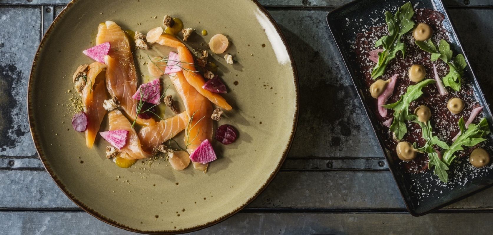 Two gourmet dishes plated with vibrant ingredients on rustic metal surface.