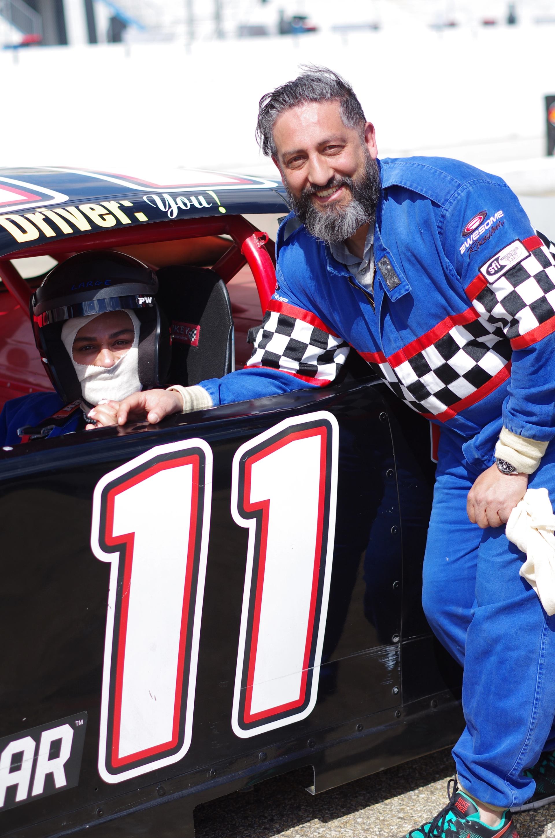 Person in a racing suit standing beside a NASCAR stock car with number 11 and another seated inside.