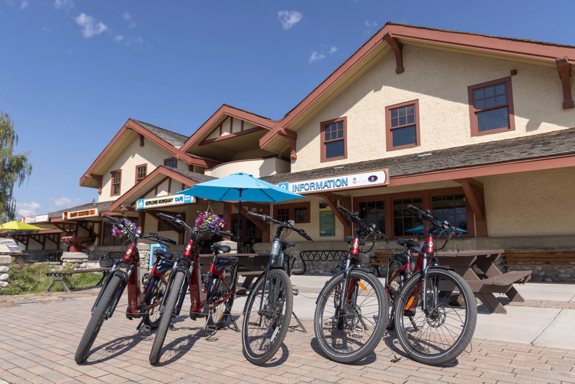 Row of parked bikes outside rustic rental shop with picnic tables and umbrella.