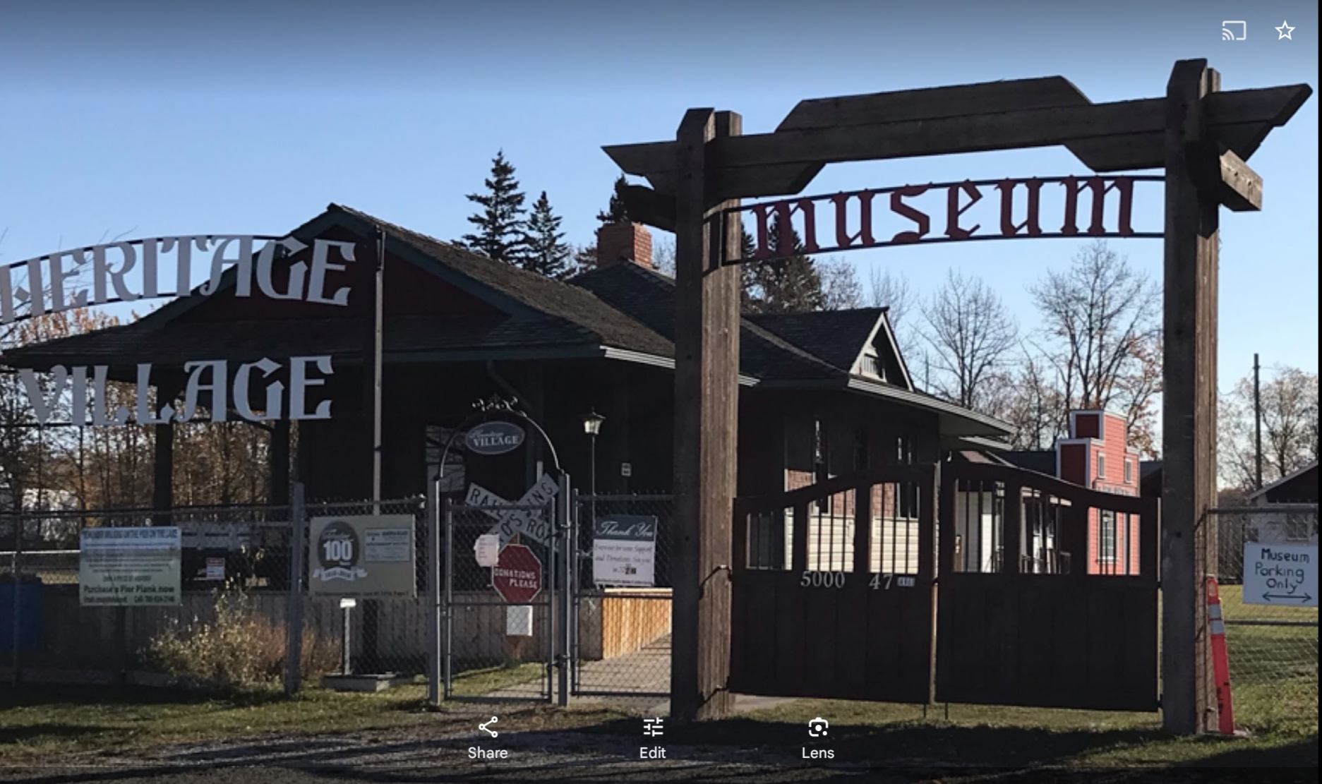 A dark-roofed building stands behind prominent entrance signs for a "Heritage Village" and "Museum."
