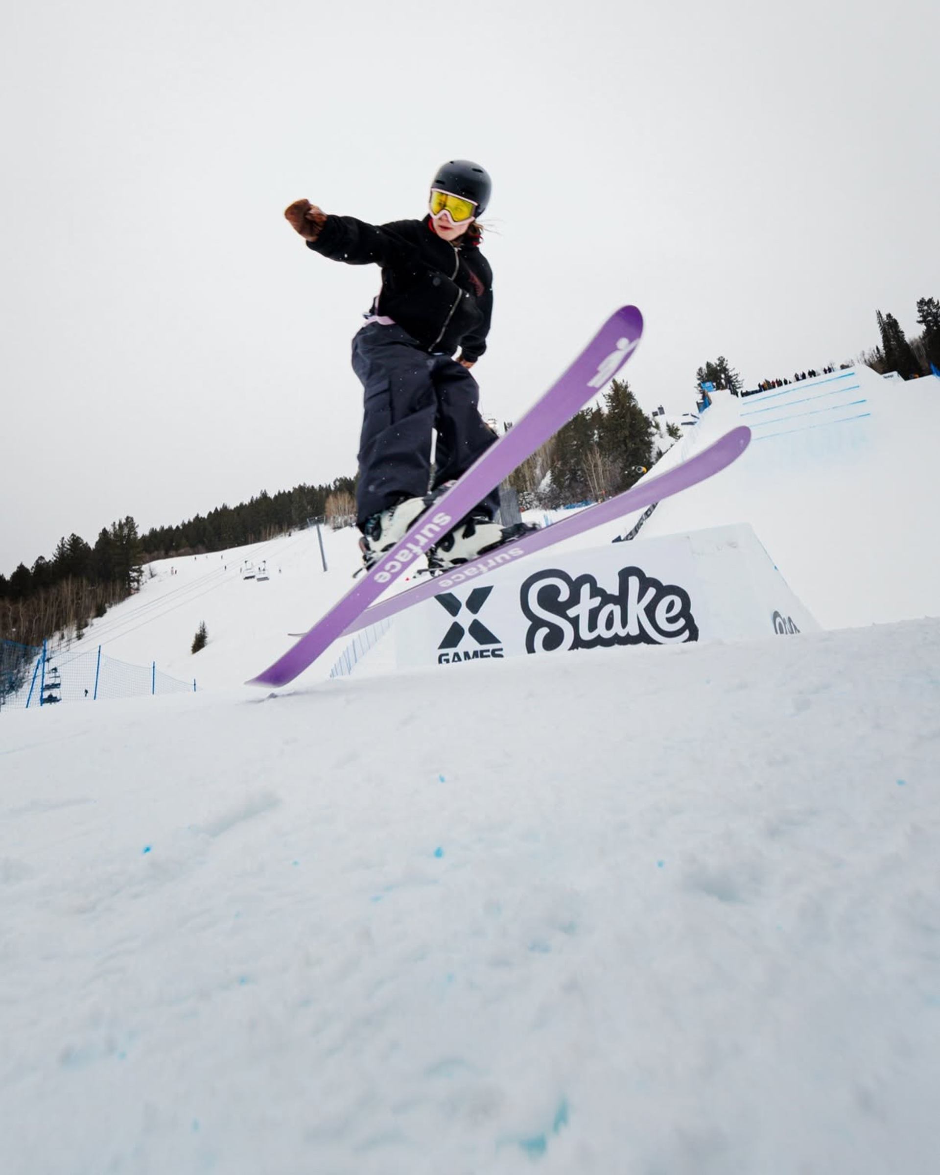 Skier wearing yellow goggles jumps over a snow park rail on purple skis.