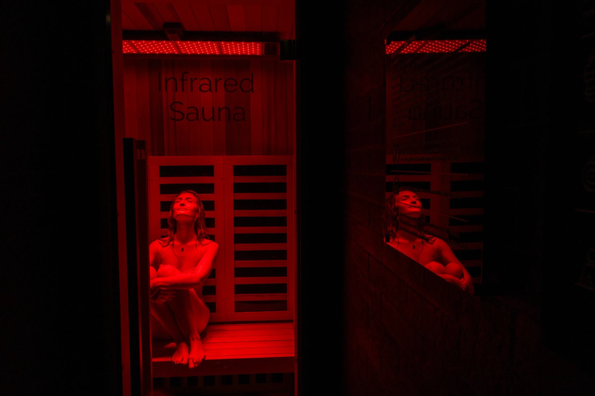Person sitting in red-lit infrared sauna, reflected in nearby mirror.