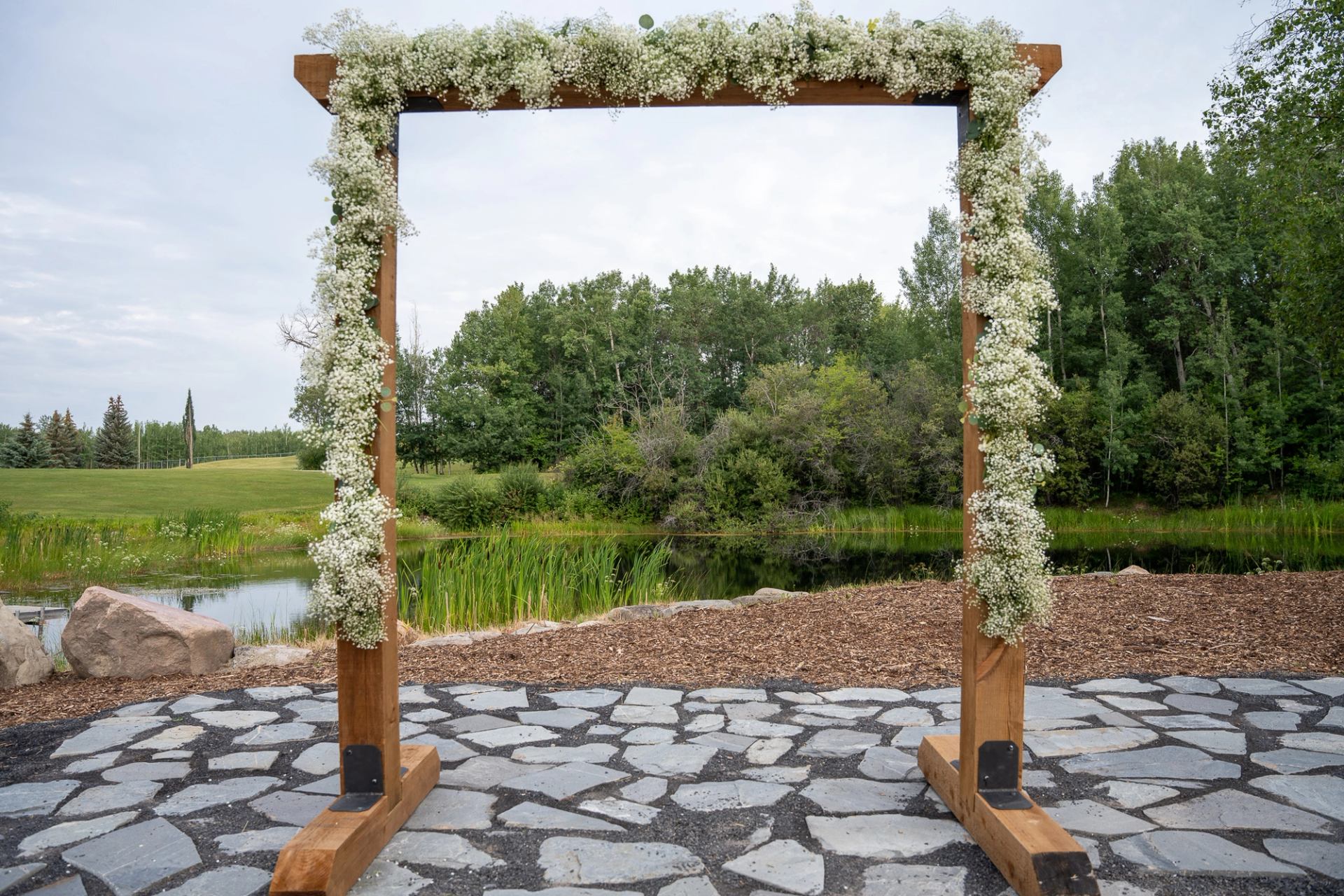 Floral arch on stone path with lush greenery and water in the background.