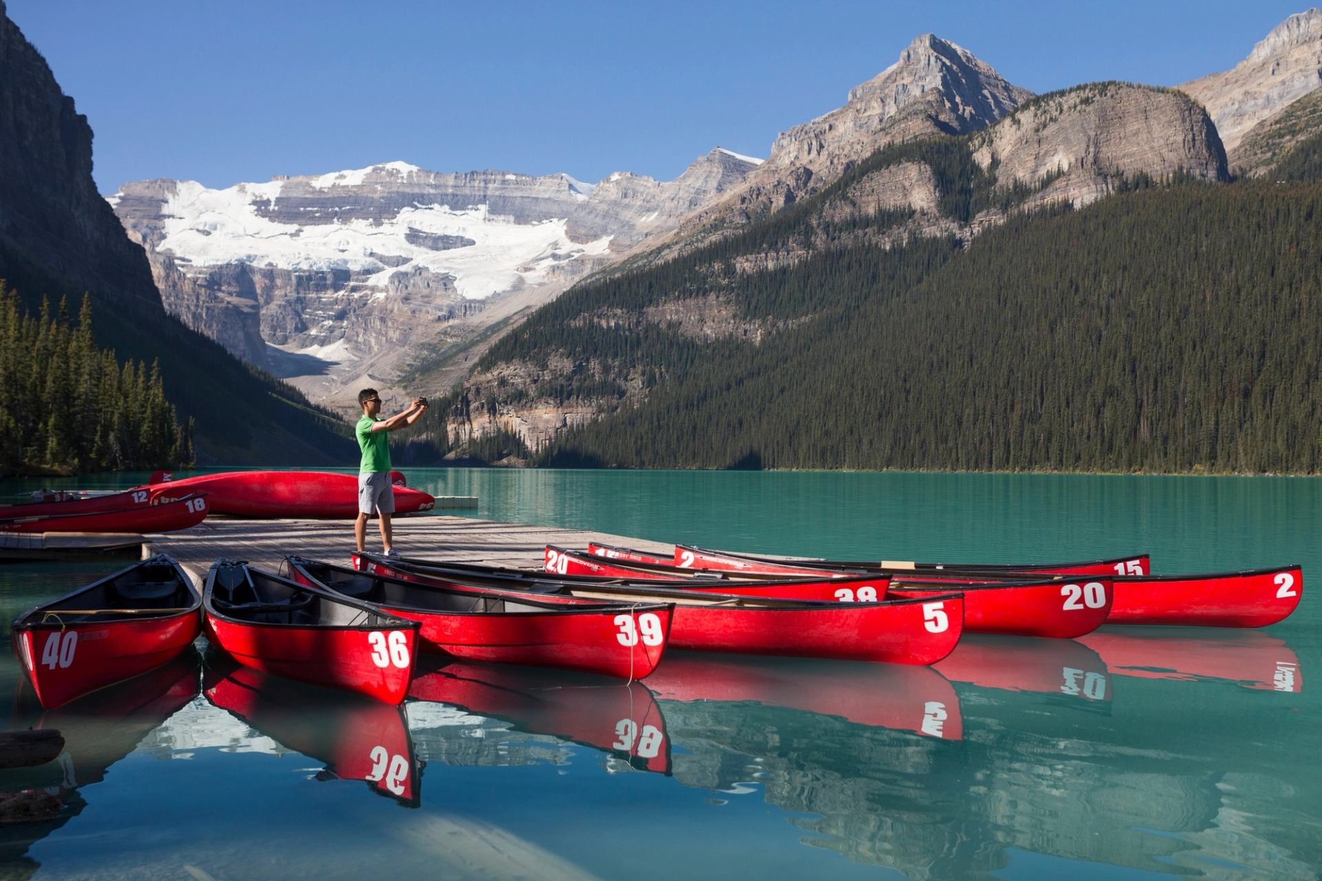 A person takes a selfie on a dock surrounded by red canoes on a turquoise lake with snow-capped mountains in the background.