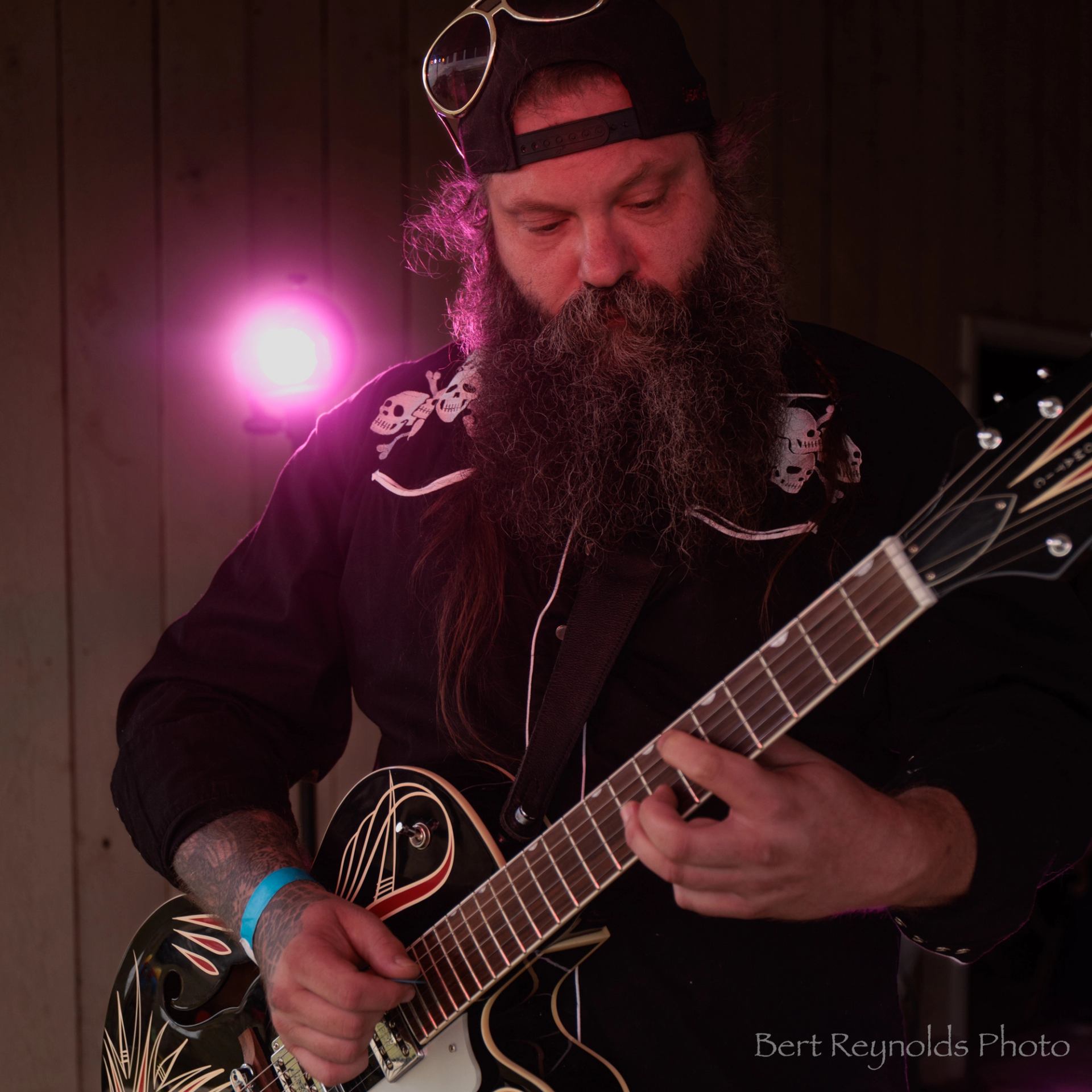 Guitarist with long beard plays decorated electric guitar under pink stage light.
