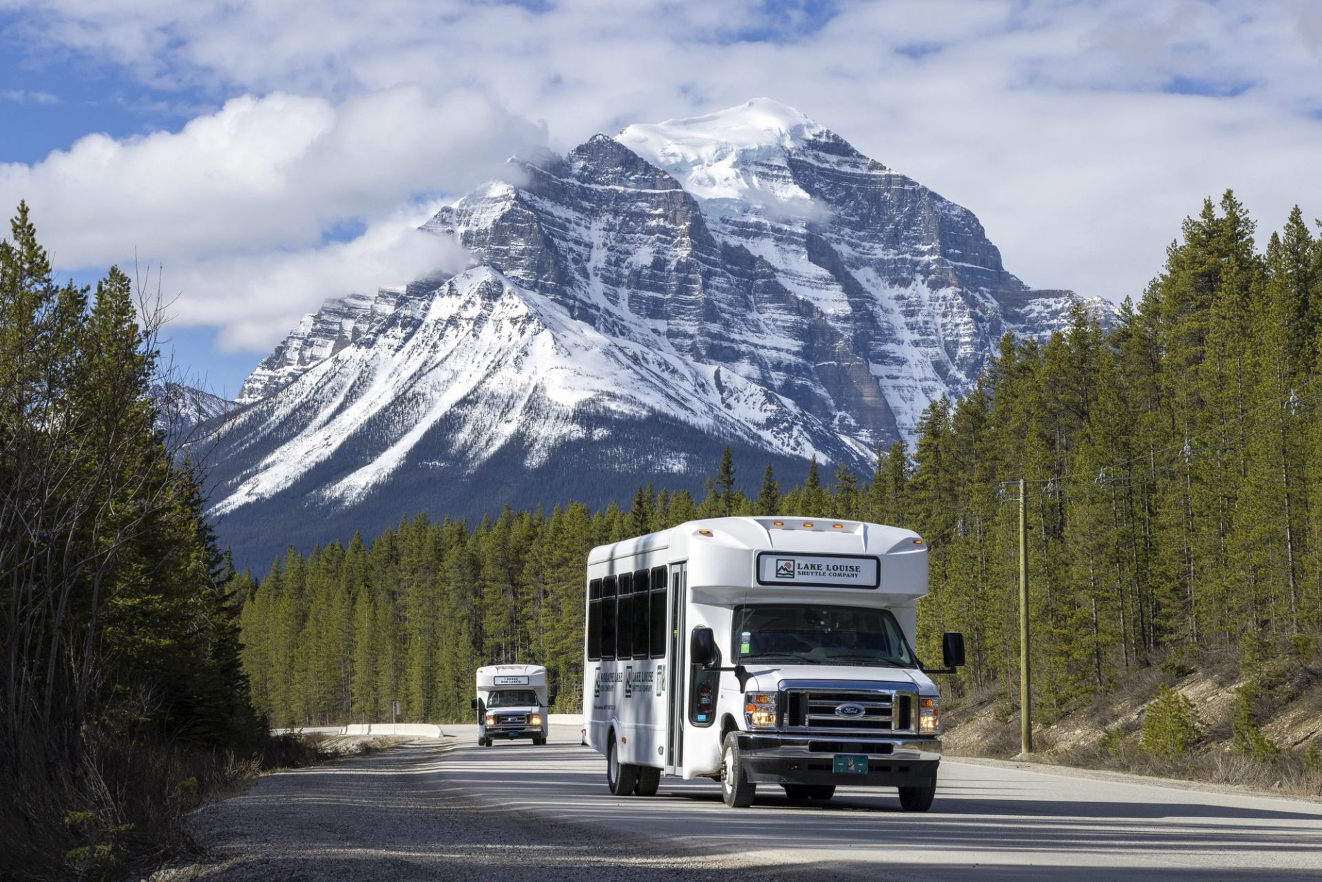 Tour buses drive along forest highway below large snow-covered mountain peak.
