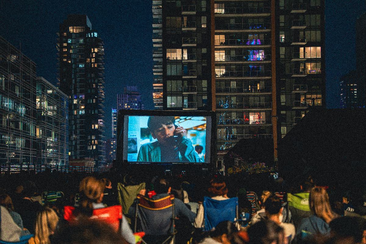 Crowd watches outdoor movie at night, seated before a large screen among tall buildings.