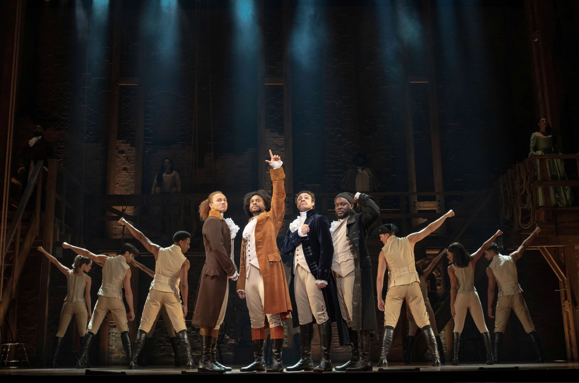 Stage performers in period costumes stand in formation under spotlights during a musical number.