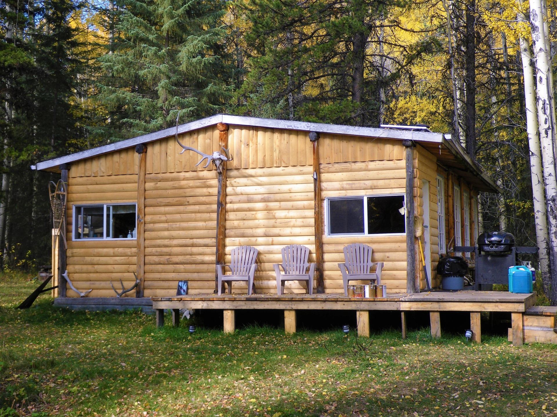 A log cabin with a deck, two chairs, grills, and surrounded by trees during autumn.