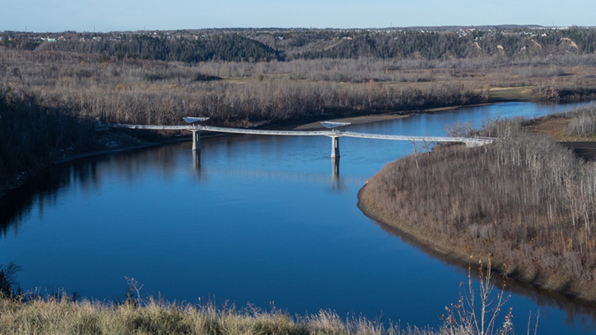 Pedestrian bridge spans a wide blue river between forested banks and distant treeline