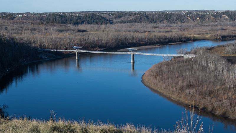 Pedestrian bridge spans a wide blue river between forested banks and distant treeline