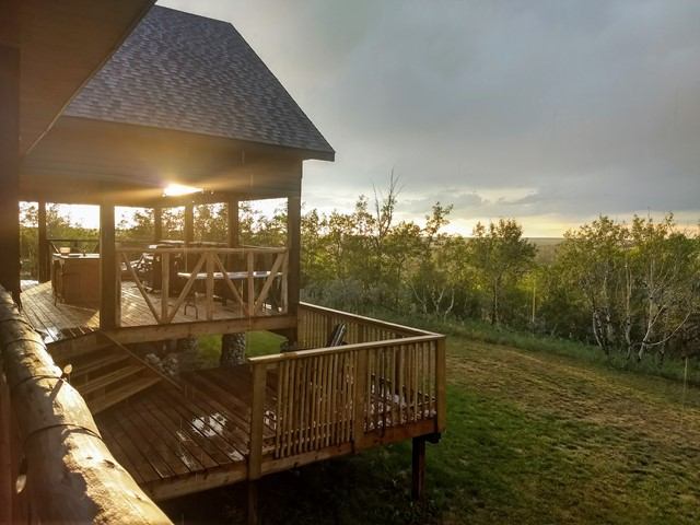 A rustic wooden cabin deck at sunrise, overlooking a grassy field and surrounded by trees. Sunlight filters through the trees, creating a tranquil ambiance.