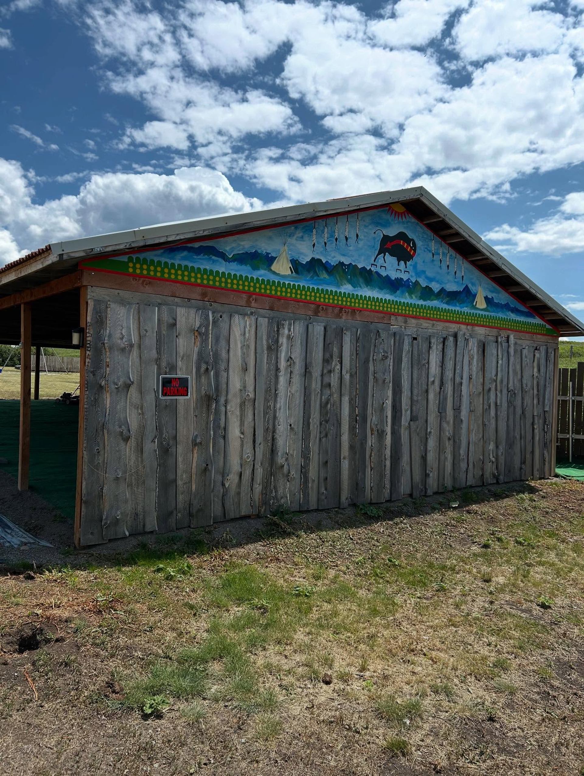 Wooden shed with painted mural of buffalo, tipis and mountains under cloudy sky.