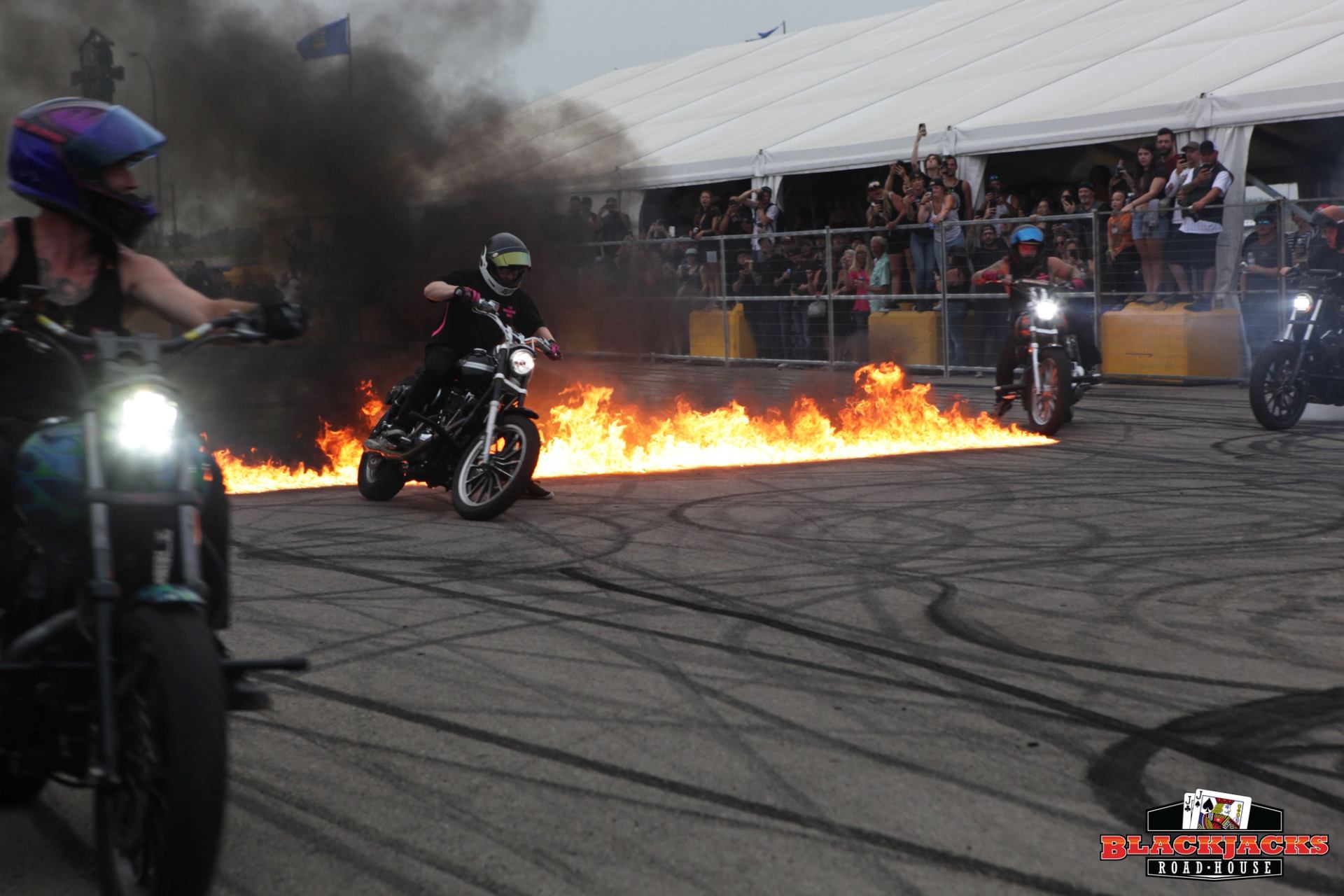 Bikers doing a burnout stunt where the rear wheel spins and creates smoke, combined with flames for dramatic effect. 