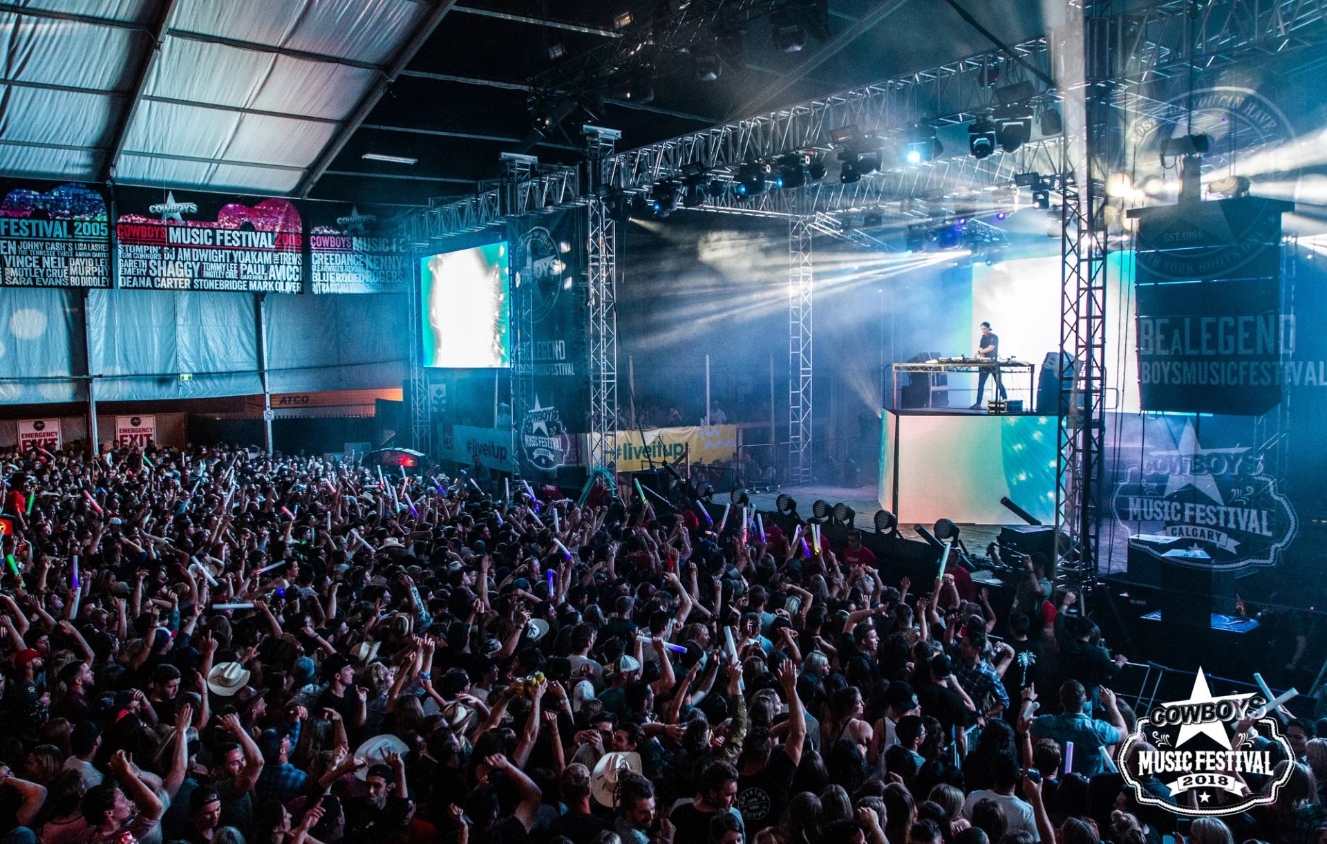 A DJ sits on a stage high above a large crowd of concert goers.