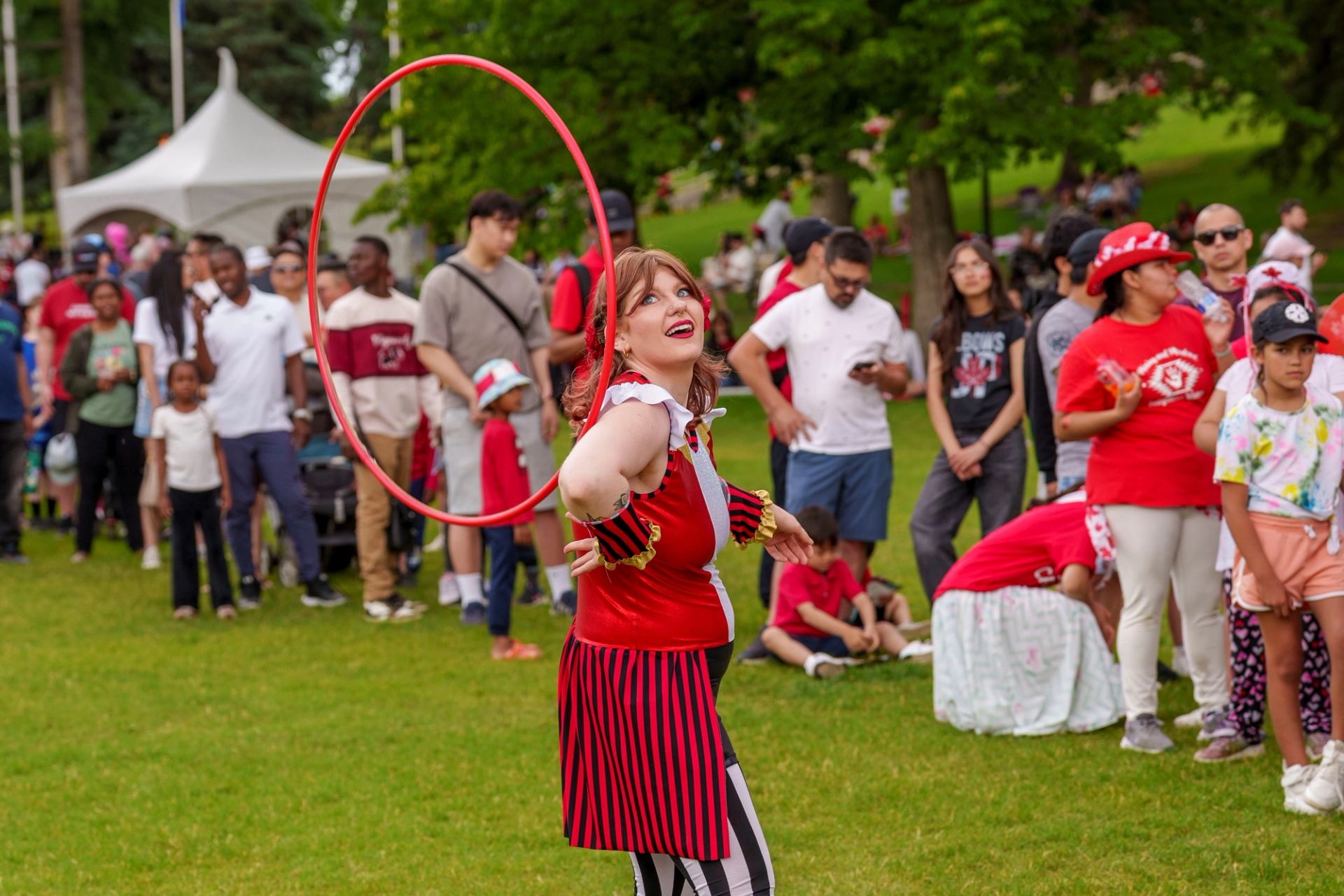 Performer in red costume spins a large hoop before a crowd in a park.