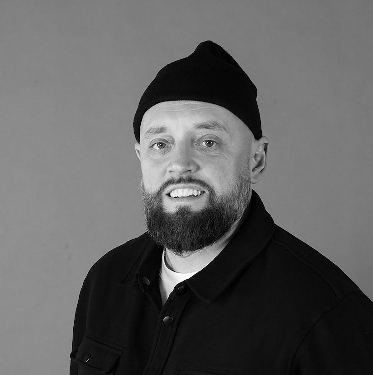 Portrait of Jerry Ropson wearing a black cap and dark jacket against a grey background.