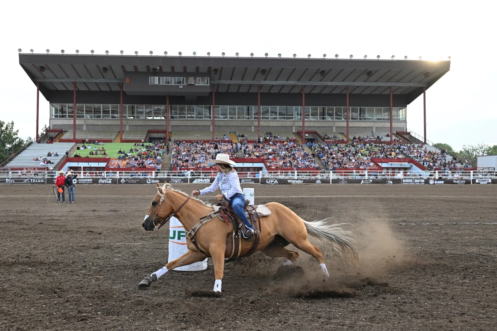 A female barrel racer on a palomino horse kicks up dirt while rounding a barrel in a rodeo arena, with a grandstand full of spectators.