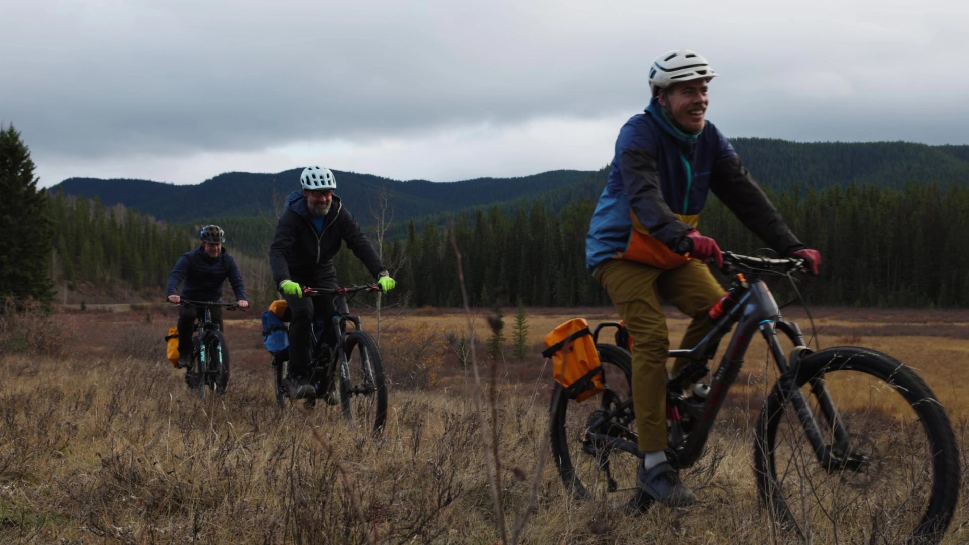 Bicyclists ride mountain bikes on a trail through a forested valley under cloudy skies.