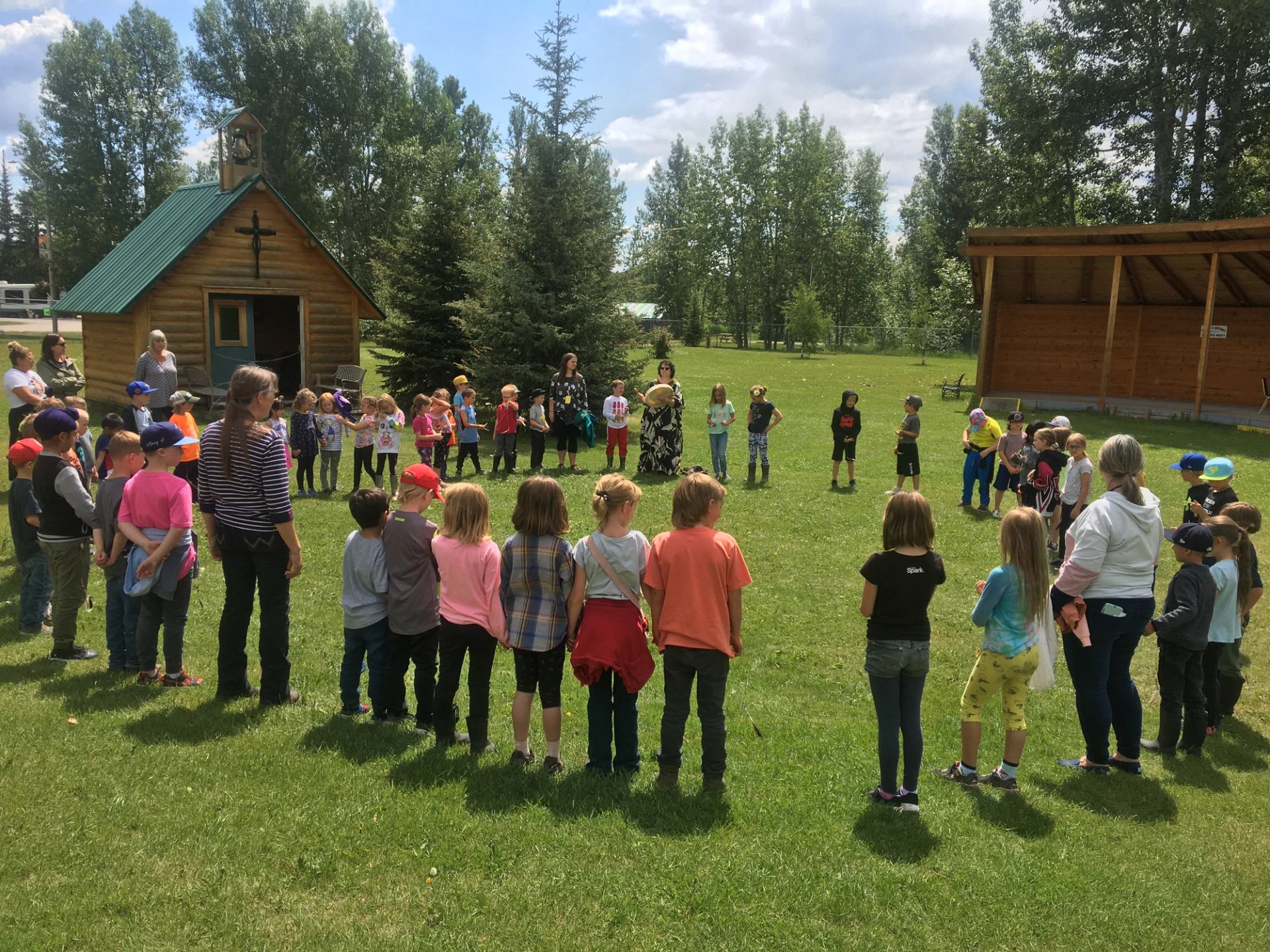 A large group of children and adult instructors stand in a wide circle on a lush green lawn during an Indigenous Peoples Day celebration. In the background, a small wooden log cabin with a green roof sits near tall trees under a bright sky.
