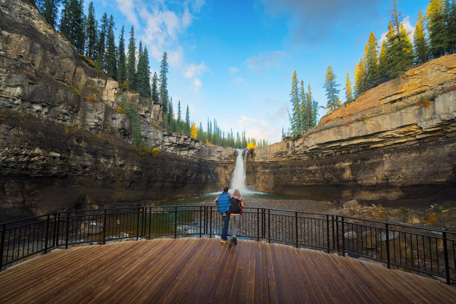 Two visitors stand on a wooden deck overlooking a waterfall in a rocky canyon surrounded by trees.