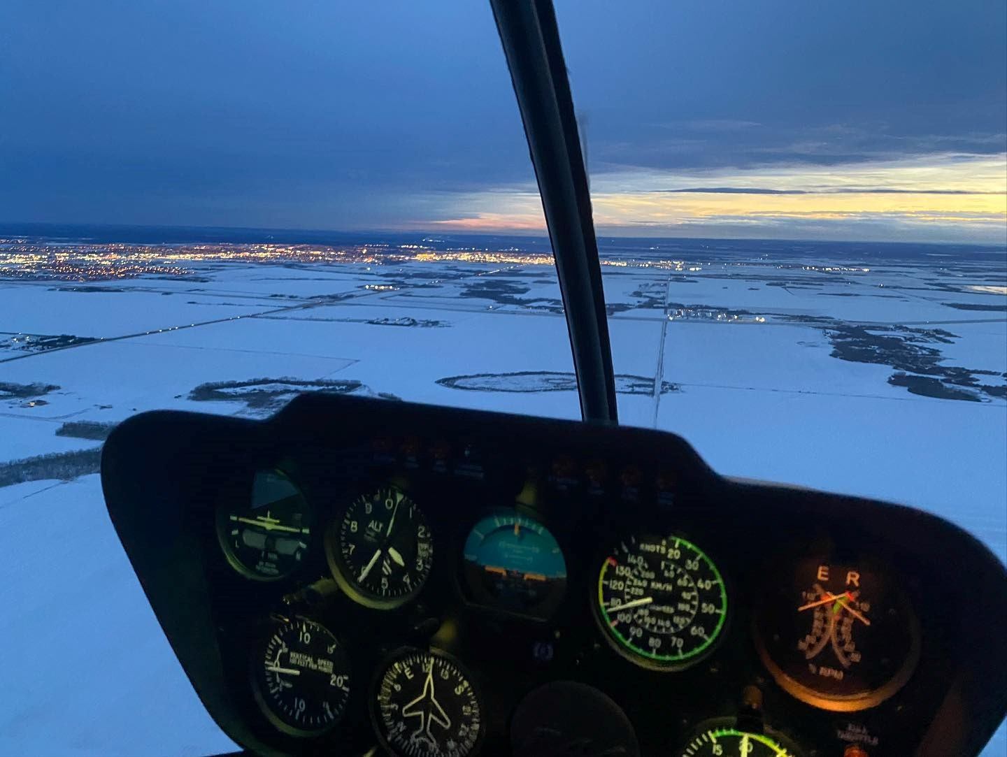 Cockpit view of helicopter flying over snowy terrain at twilight.