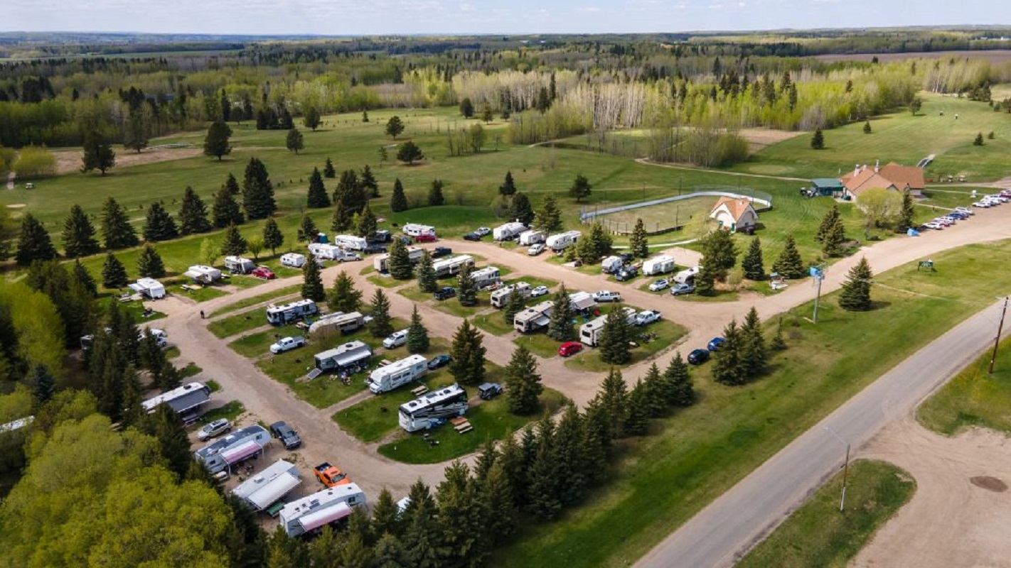 Aerial view of RV campground with trailers, trees, dirt roads and surrounding open fields.