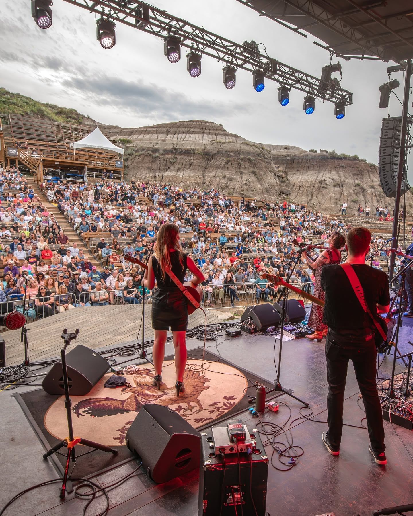 Outdoor concert stage with band performing to large seated crowd before rocky cliffs.