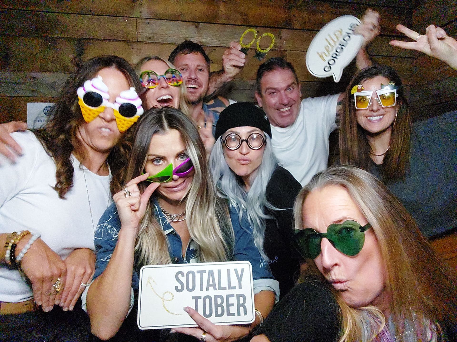 Eight people in novelty glasses pose with playful signs in front of a wooden wall.