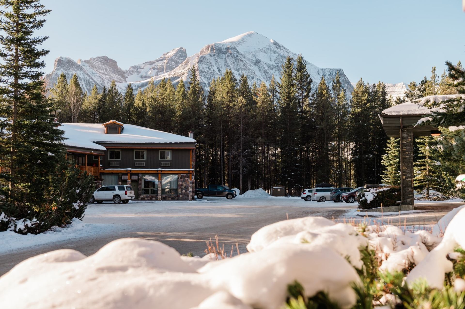 Snow-covered lodge with parked cars and towering mountains in the background.