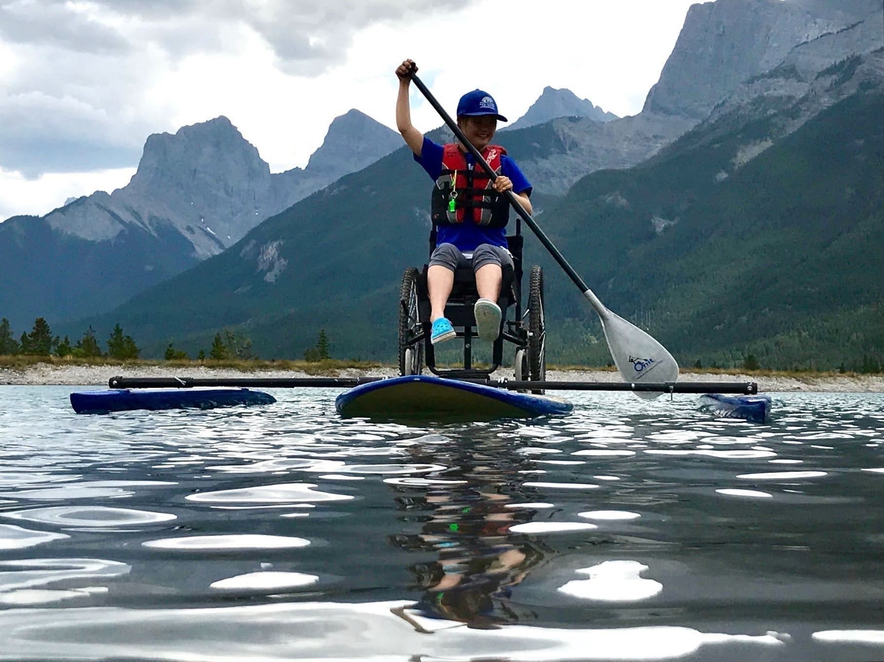 Person in wheelchair kayak paddling on calm lake with mountain view.