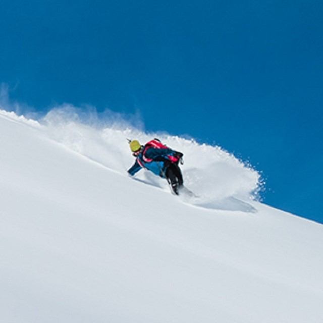 Skier in colorful gear glides down snowy slope under a bright blue sky.