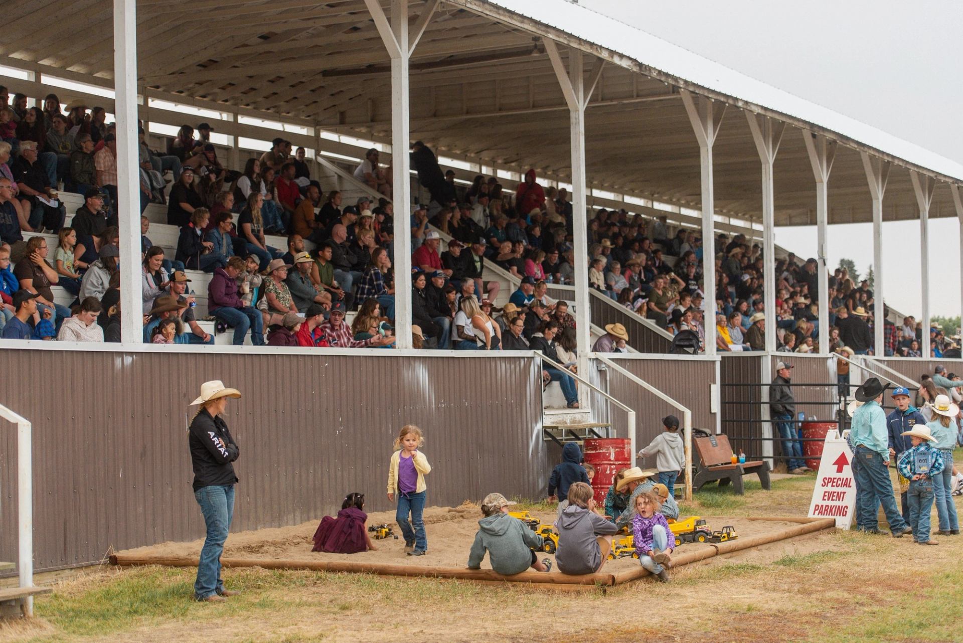 Crowd seated in a rodeo grandstand under white steel rafters, with children in front.