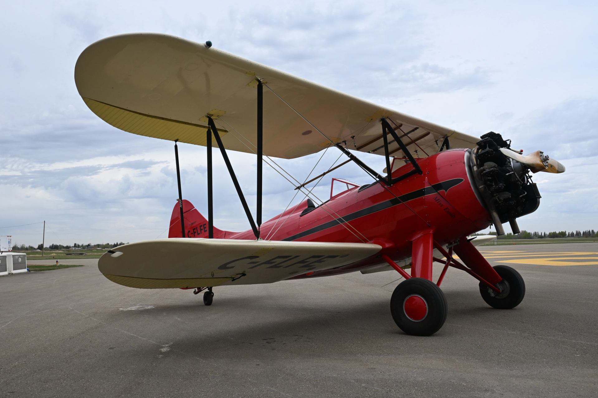 Red vintage biplane with cream wings parked on airport tarmac under cloudy sky.