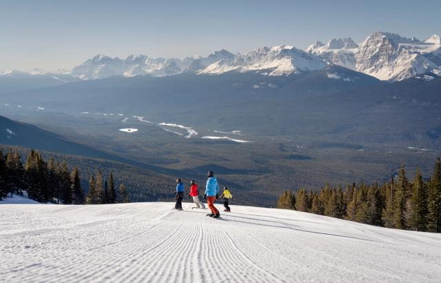 A group of four snowboarders carve on an open groomed run with mountain views while snowboarding at Lake Louise Ski Resort in Banff National Park