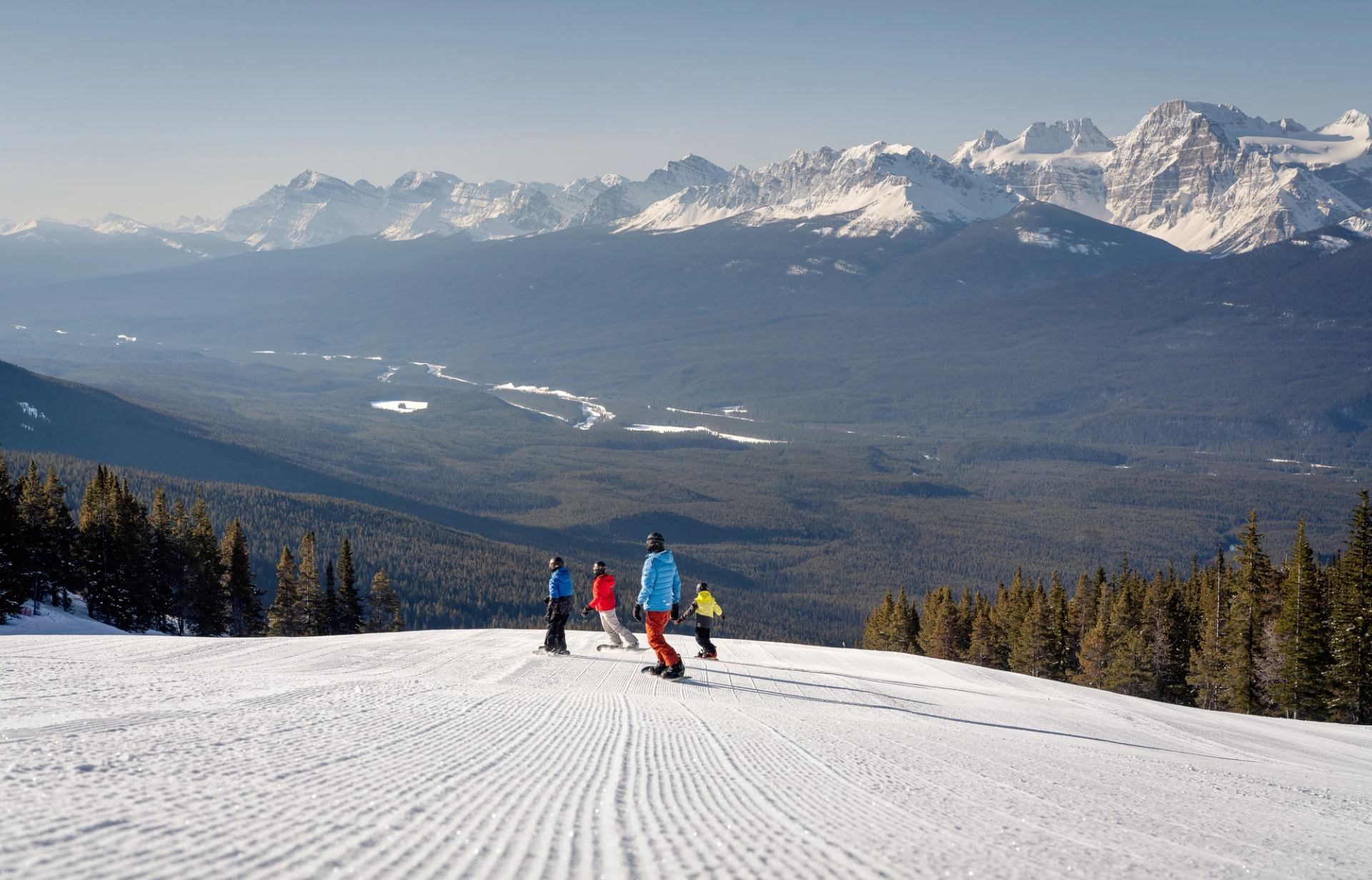 Four snowboarders carve down groomed run with mountain views at Lake Louise Ski Resort.