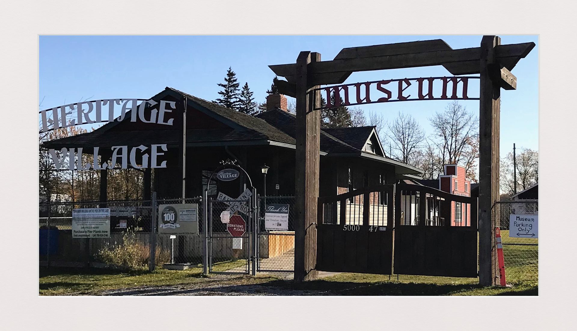 Heritage Village museum entrance with wooden gate, fence, signs and surrounding trees.