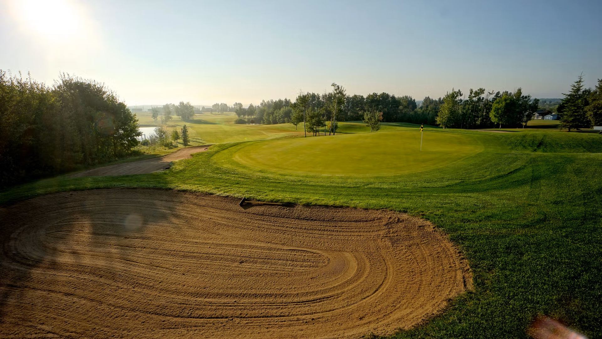 Golf course green with sand bunker in foreground and trees lining fairway.