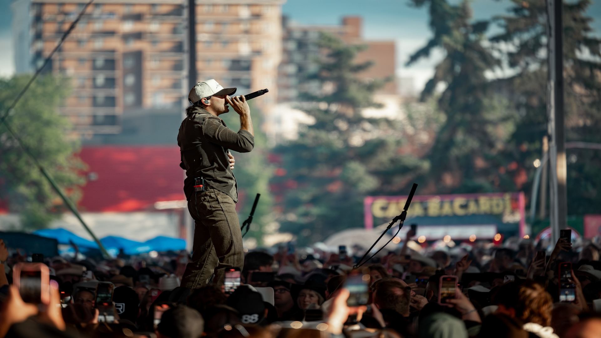 Performer onstage singing to a large outdoor crowd with phones raised in the air.