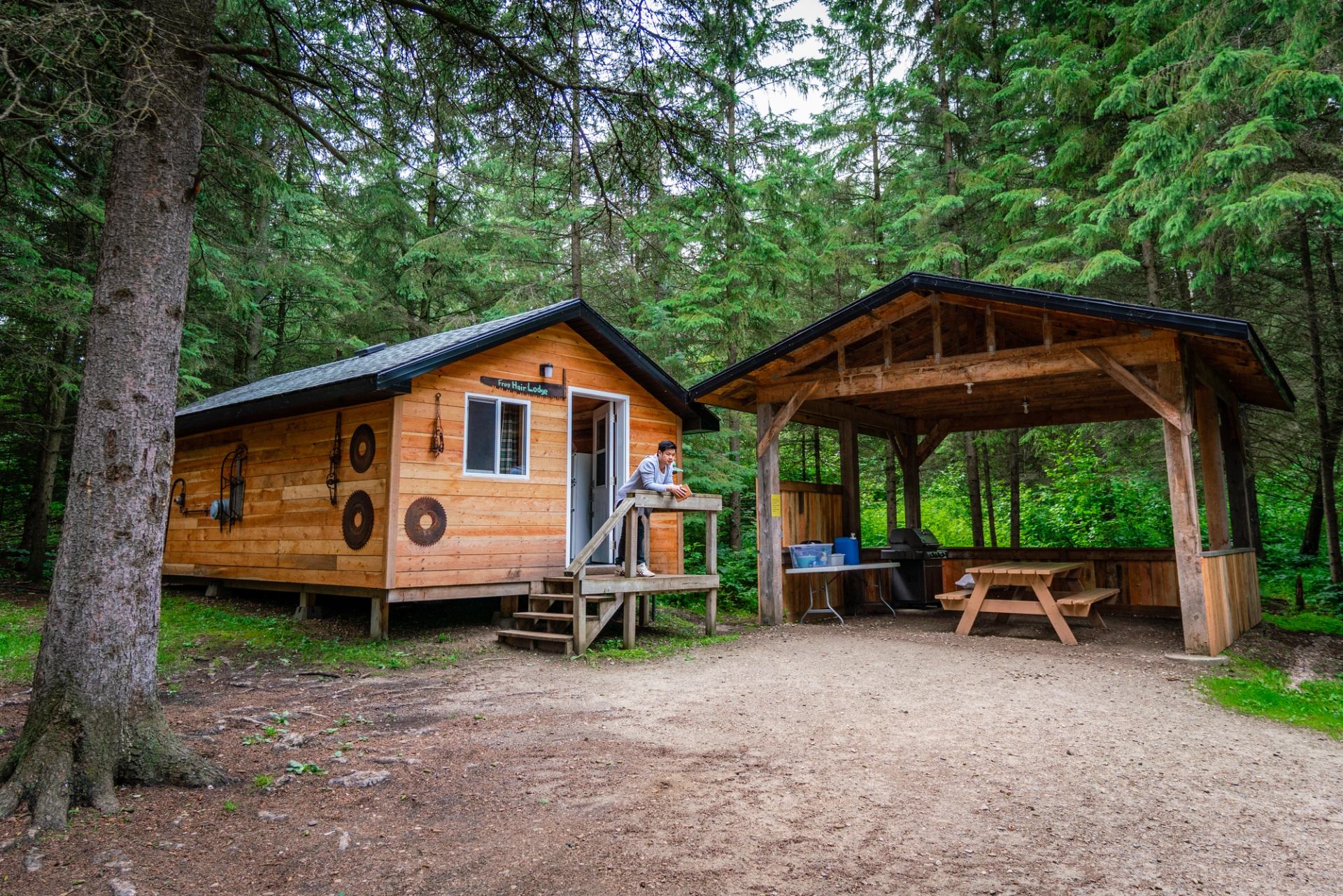 A person relaxes outside a cabin in the woods, next to a covered picnic area."