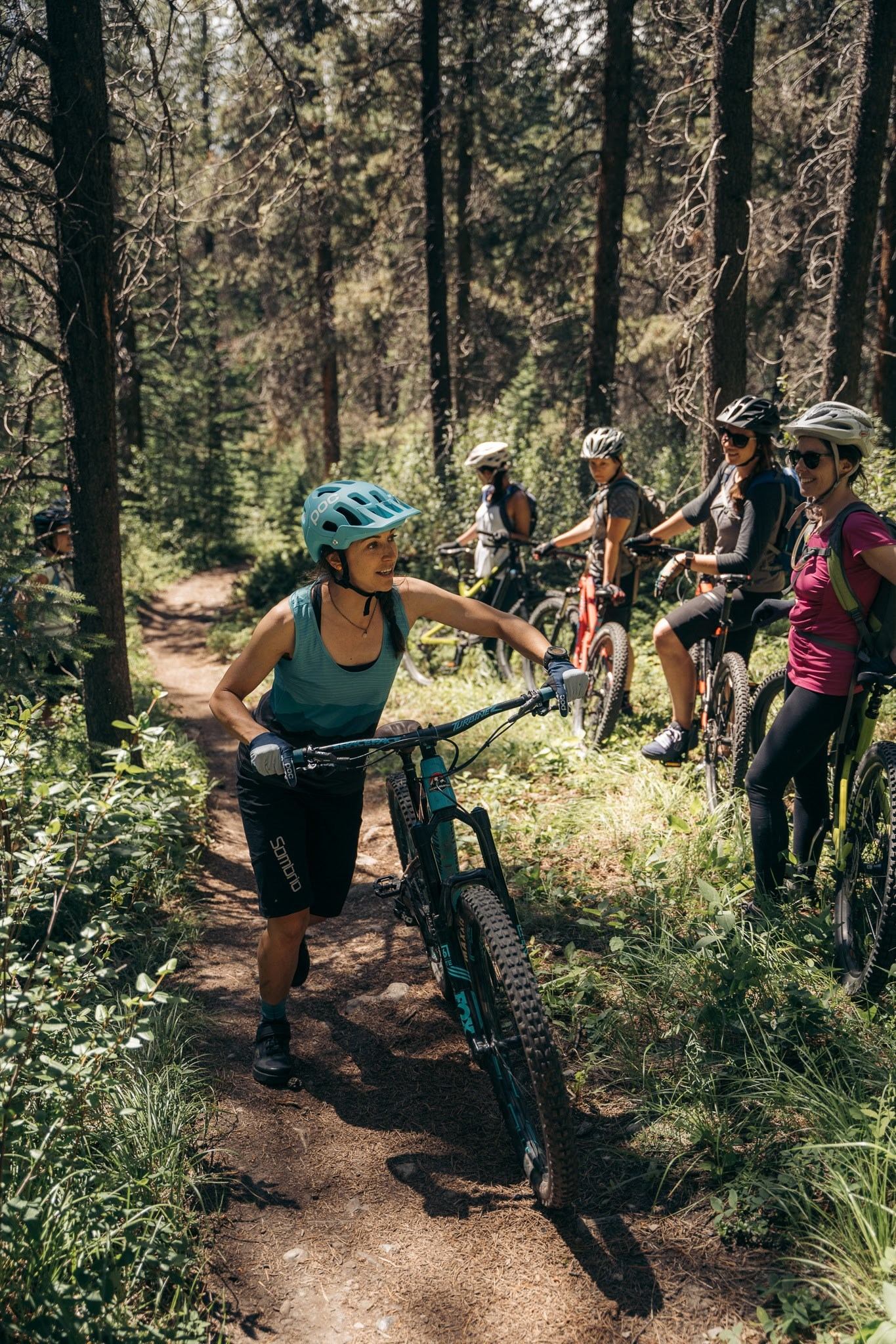 A group of cyclists pause on a sunlit forest trail; a woman in front holds her bike, looking back joyfully.