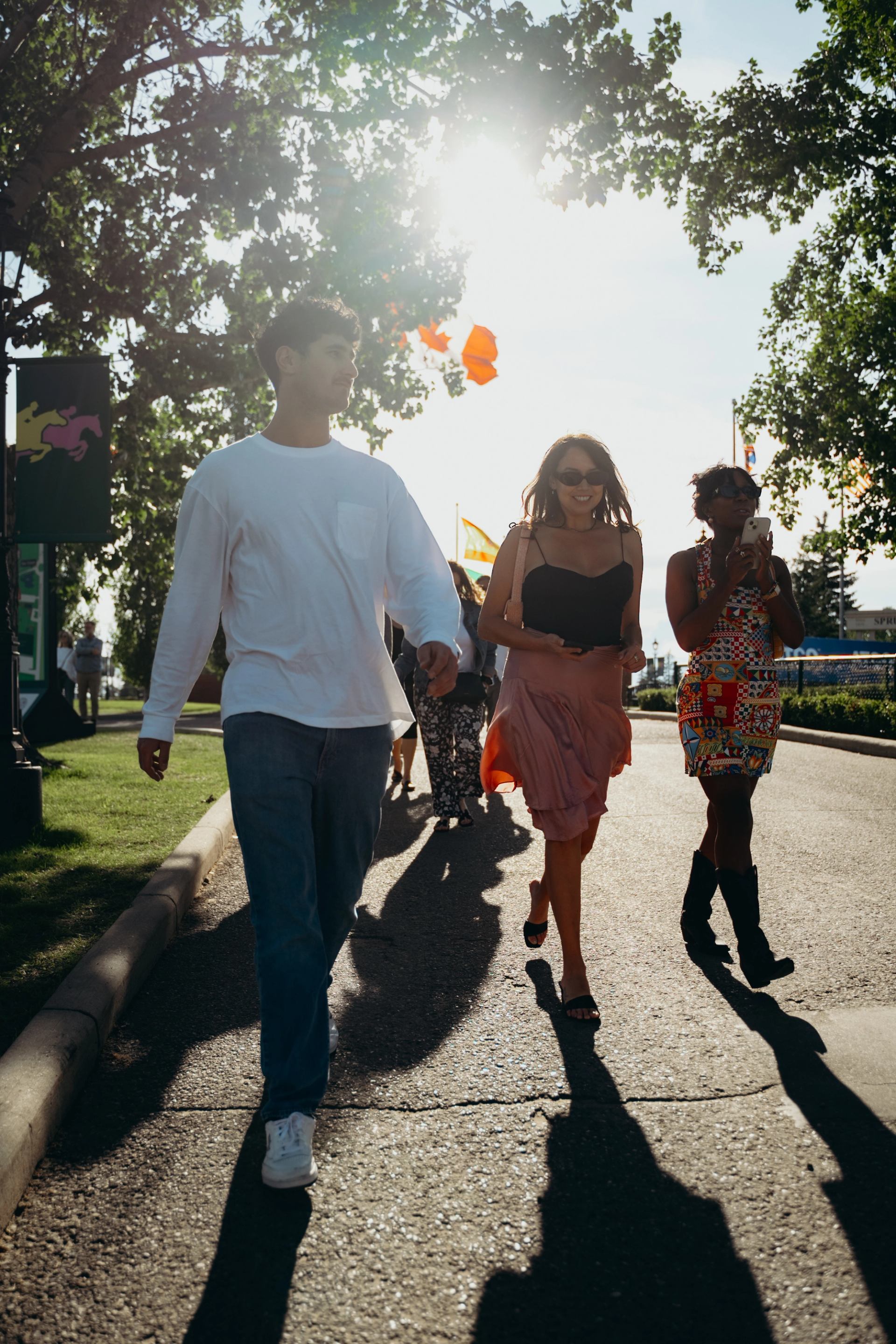 Group of friends walk at a sunny event with flags, trees, and shadows in the background.