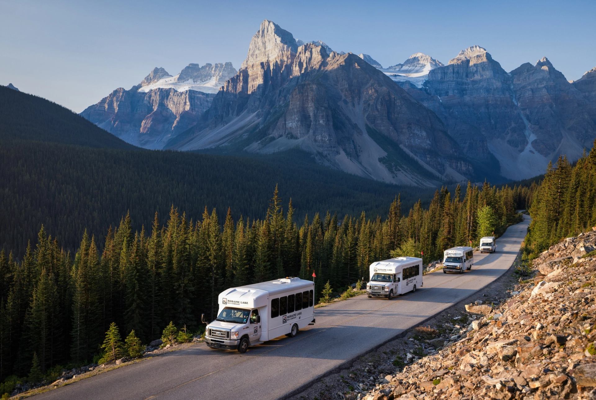 Moraine Lake Bus Company shuttles drive through forested mountain road under rocky alpine peaks.