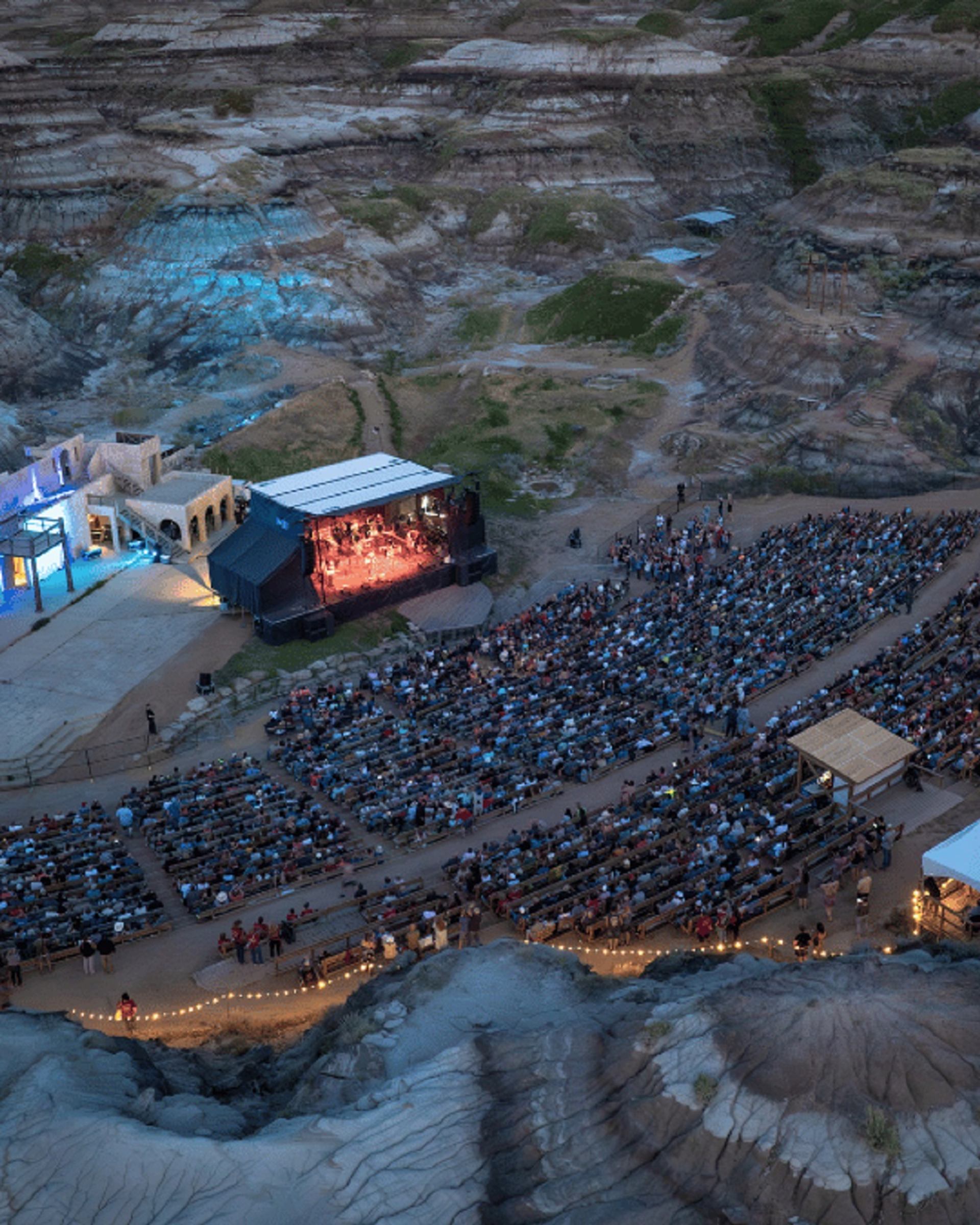 Outdoor amphitheatre in rocky canyon with large audience watching illuminated stage performance.