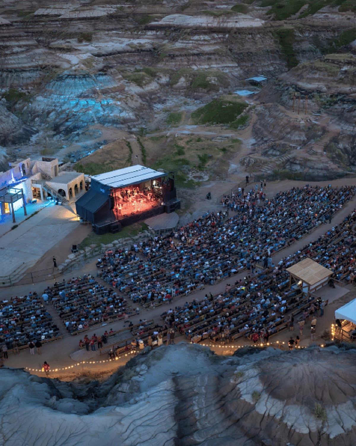 Outdoor amphitheatre in rocky canyon with large audience watching illuminated stage performance.