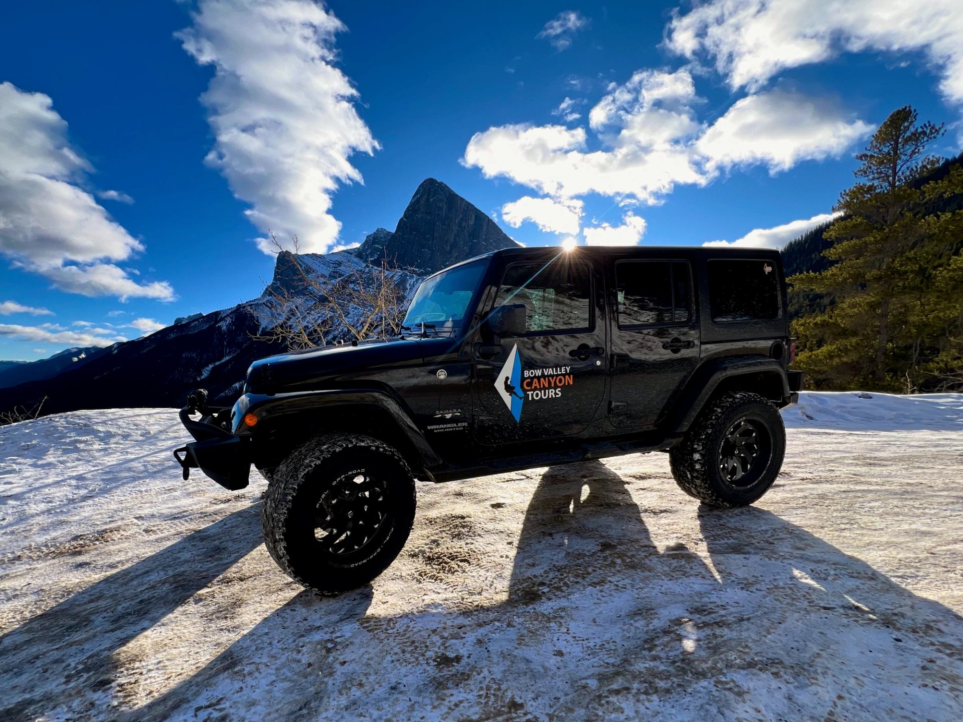 Black Jeep on snowy ground with dramatic mountains and bright blue sky in the background.