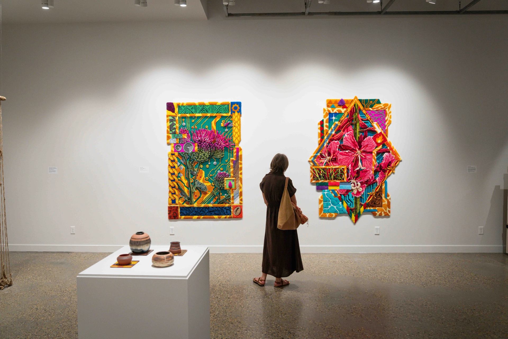 Gallery visitor viewing two colourful floral textile artworks beside a pedestal of ceramic vessels.
