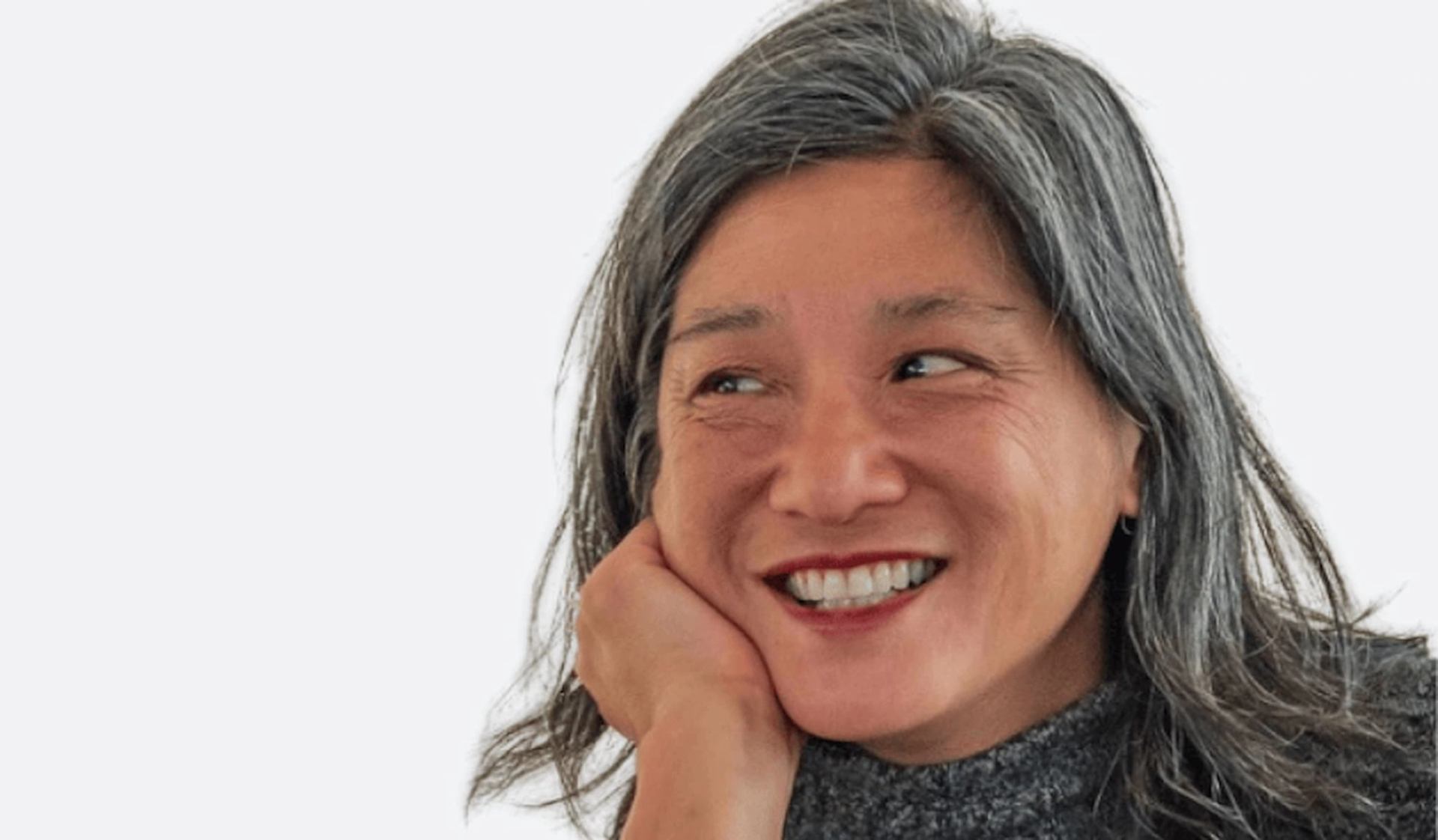 Person with long greying hair rests chin on hand, looking sideways against plain background.