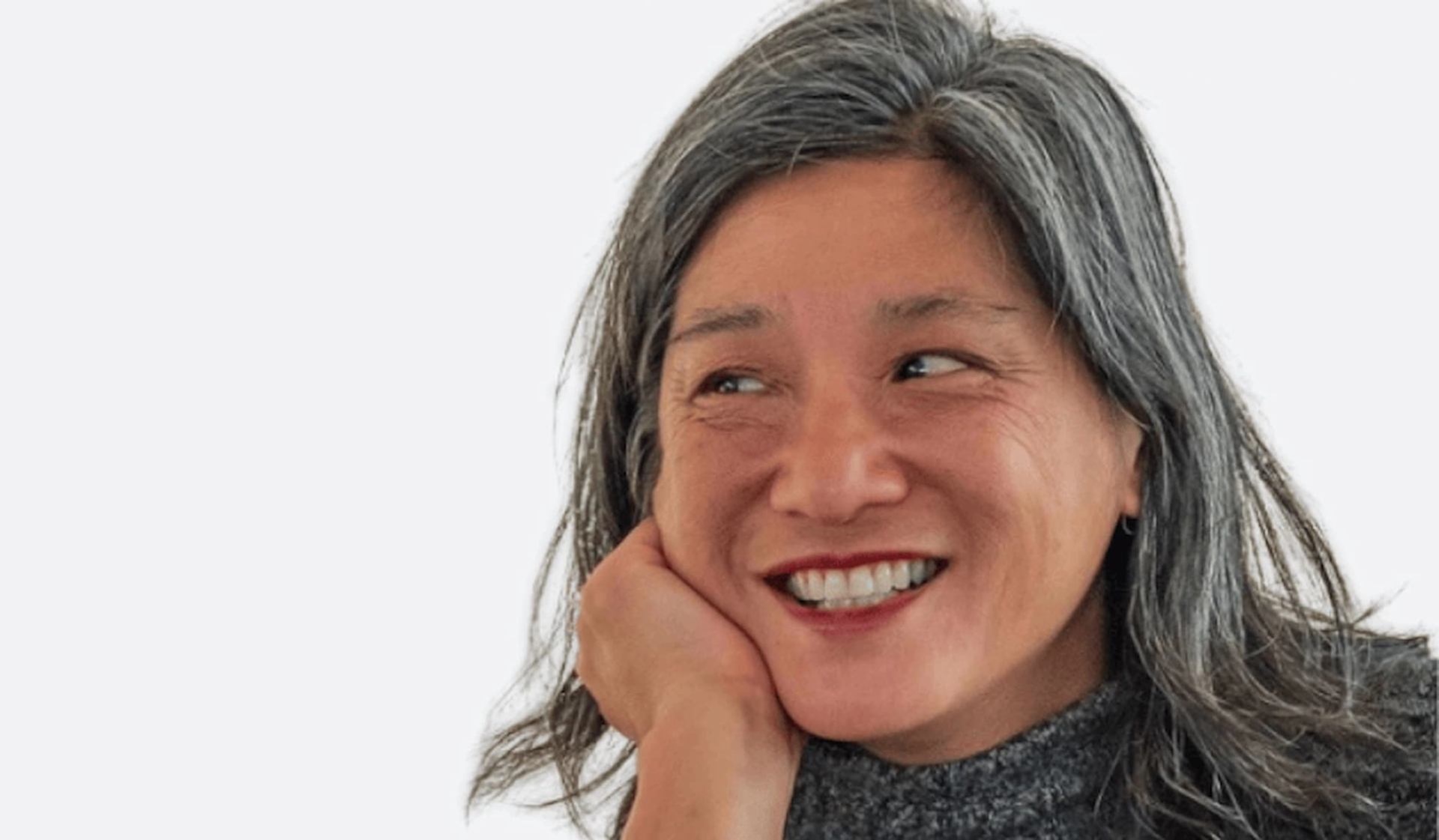 Person with long greying hair rests chin on hand, looking sideways against plain background.