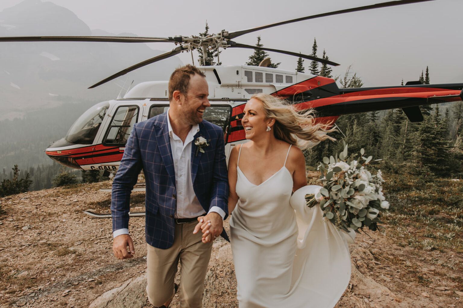 Two people in wedding attire holding hands near a helicopter on a mountain landing spot.